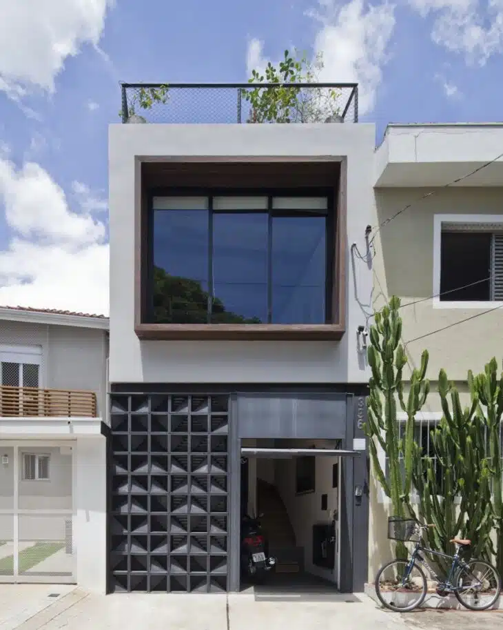 Fachada de casa simples em terreno estreito Fachada estreita de residência compacta com janela ampla, elementos vazados e vegetação junto à entrada