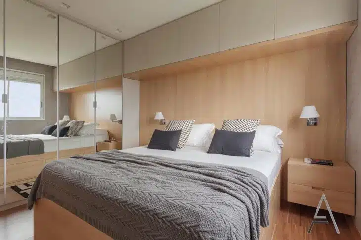 Quarto com cama integrada a painel de madeira, armários superiores e portas de espelho