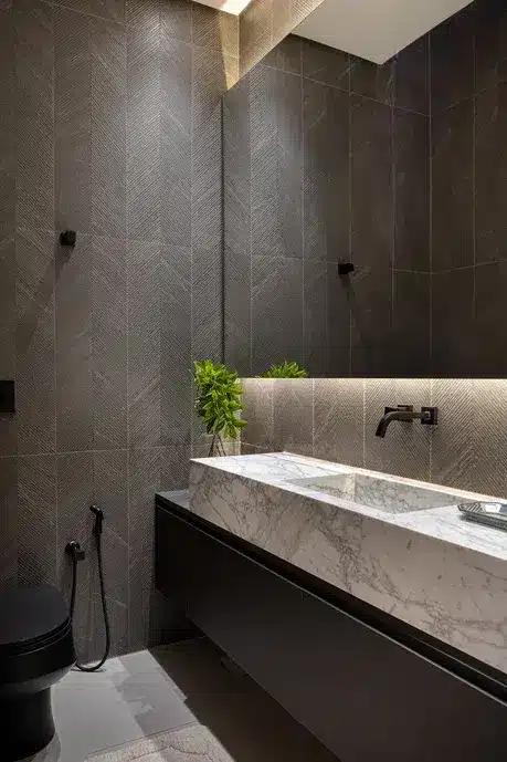 Você sabia que a paginação escama e a espinha de peixe podem transformar seu espaço? Descubra as diferenças e inspirações! 2 Banheiro com foco de iluminação na parte da pia embaixo do espelho. Uma planta na decoração. Paredes com revestimento em tons cinzas.