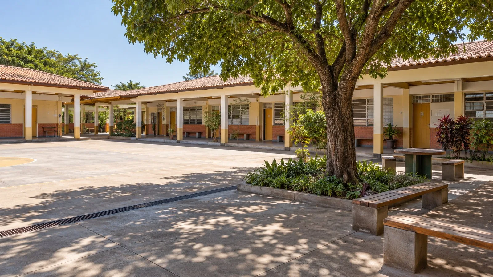 Arquitetura escolar: como planejar espaços de ensino 4 Pátio escolar com área pavimentada livre, bancos à sombra de árvore e circulação coberta junto às salas.
